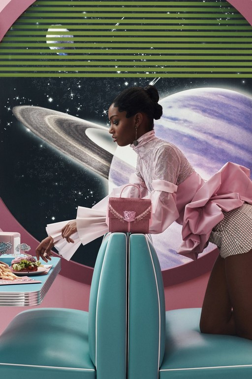 Glamorous Black female model poses in a retro-futuristic diner setting with space scene backdrop, showcasing a pink handbag.