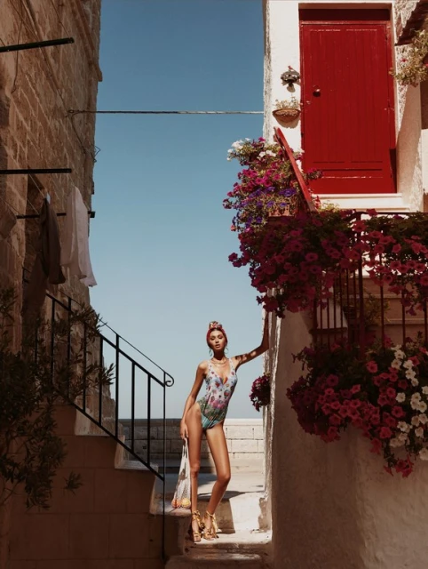 Woman in floral swimsuit poses on steps of European building, with red door, flowers, and blue sky backdrop. Fashion editorial.
