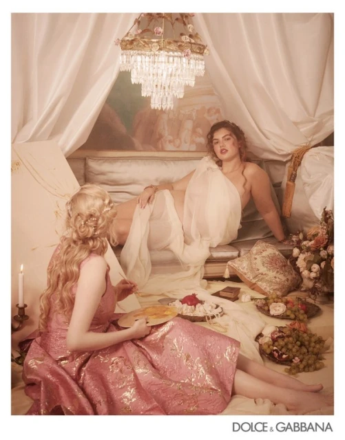 Two women in classical fashion pose in a bedchamber with an ornate chandelier, painting, and fruit. Elegant and artistic atmosphere.