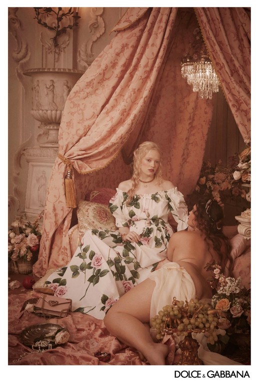 Two women pose in a luxurious indoor setting with draped textiles, flowers, and a chandelier, Dolce & Gabbana logo at the bottom.