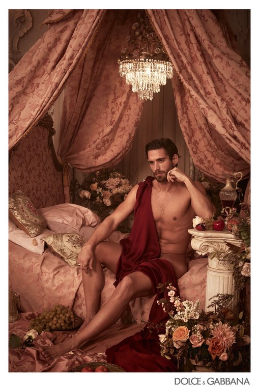 Fashion image with a male model draped in red fabric sitting on a bed surrounded by flowers, fruit, and a chandelier in a bedroom setting.