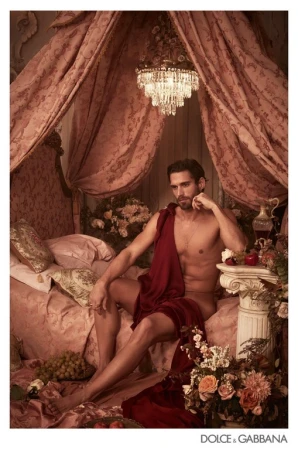 Fashion image with a male model draped in red fabric sitting on a bed surrounded by flowers, fruit, and a chandelier in a bedroom setting.