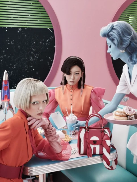 Three models at a retro diner table with a pink and turquoise color scheme, featuring a purse, milkshake, lava lamp, and donuts.