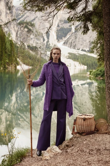 Fashion editorial shot of a woman in a purple suit holding a fishing net, posing in front of a lake and mountain range on an overcast day.