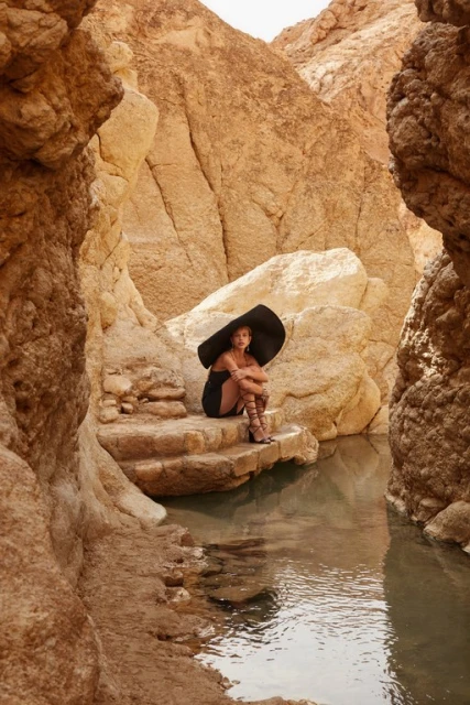 Woman in a black dress and large hat sits on rock formations next to a pool of water surrounded by a canyon with rock walls.