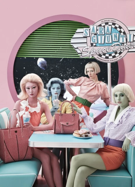 Four women with bold makeup and retro hairstyles pose at a diner with pink and turquoise decor, showcasing purses and donuts.