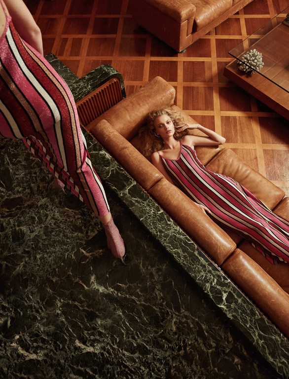 Two women in striped jumpsuits pose in an indoor setting, one reclining on a brown leather sofa with a green marble table nearby.