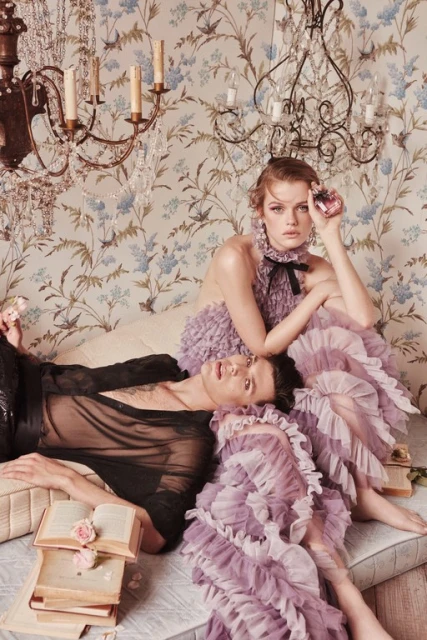 Indoor fashion portrait of a woman in a ruffled purple gown sitting next to a man lying on a bed covered in books.