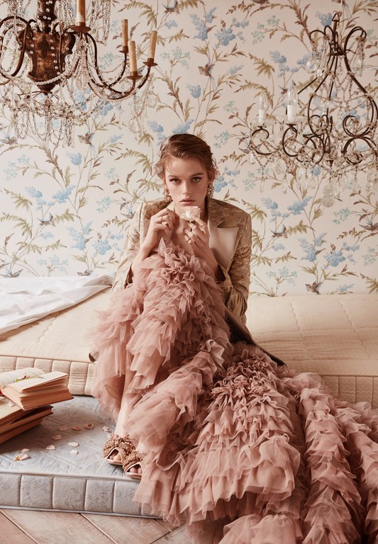 Fashion shot: Woman in a formal jacket and pink ruffled dress sitting on a bed with books, under a chandelier with floral wallpaper.