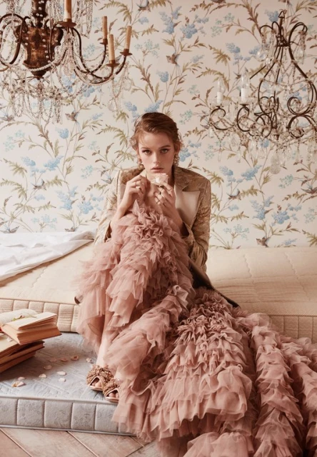 Fashion shot: Woman in a formal jacket and pink ruffled dress sitting on a bed with books, under a chandelier with floral wallpaper.