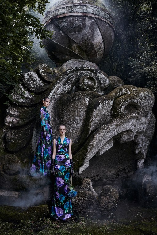 Two women in floral gowns pose near a weathered stone sculpture in a mysterious outdoor setting with fog. Fashion editorial.