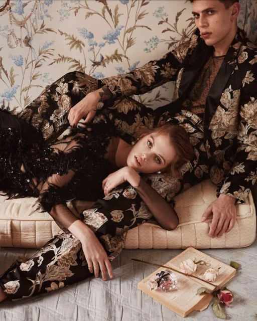 Editorial fashion shot of a stylishly dressed couple lounging on a floral chaise lounge with a book and rose in a soft light.