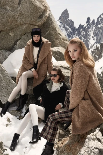 Three women in stylish winter coats pose on rocky, snowy mountain terrain with distant peaks, creating a fashion-forward outdoor scene.