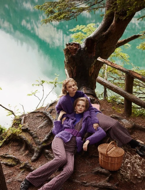 Two blonde female models in purple knitwear and striped pants pose on a tree trunk by a lake, with a wicker basket nearby.