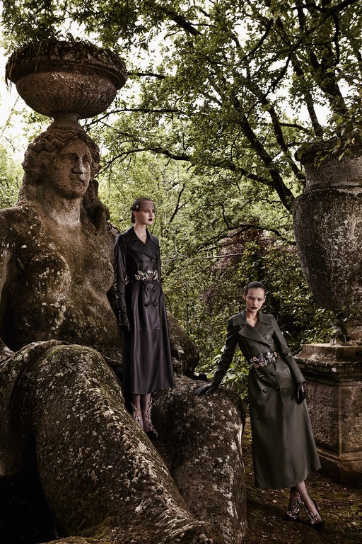 Two fashion models pose in long coats next to a mossy stone statue in a lush green park with trees in the background.
