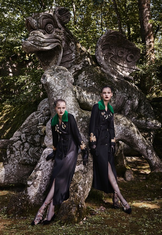 Two fashion models pose in black coats beside a large stone dragon statue in a wooded garden on an overcast day in a stylish outdoor fashion shoot.