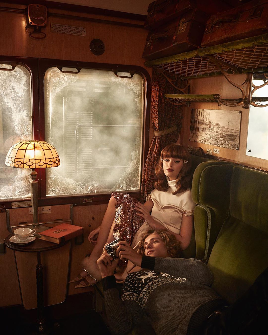 Vintage train scene: a woman in a dress sits beside a man with a camera in an antique train car with luggage racks and frosted windows.
