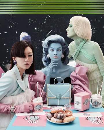 Three models pose with pastel makeup, wigs, and outfits at a table with pastries, a purse, and cups against a space backdrop in a studio.