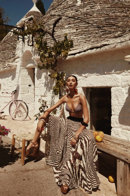 An adult woman wearing a bikini top and skirt sits on a bench outside white stone buildings with a bike in the background.