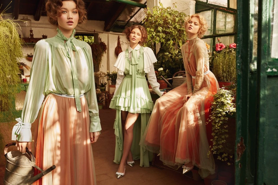 Three women in pastel gowns pose in a sunlit greenhouse-like space surrounded by plants and vintage gardening tools, evoking a vintage fashion editorial.