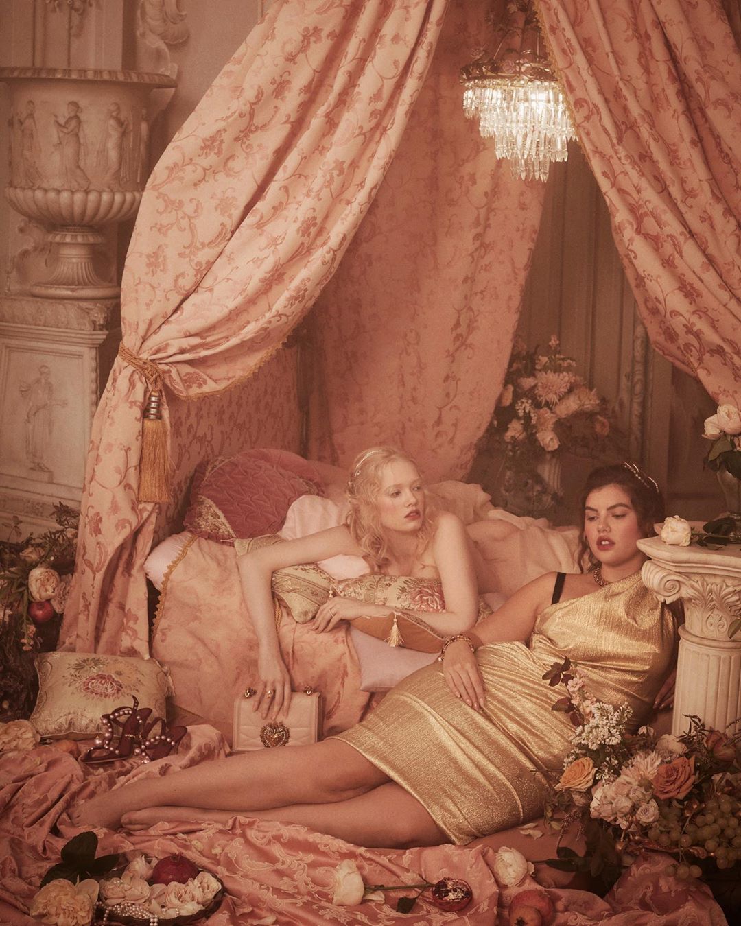 Two women lounge on a bed in a boudoir setting with vintage styling, draped in pink fabrics, surrounded by fruit, flowers, and classic decor.