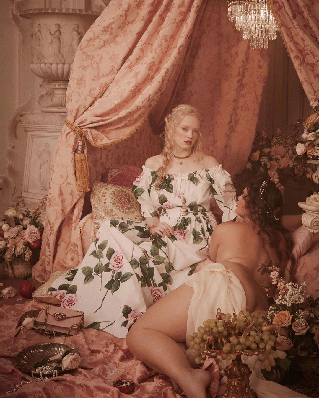 Two women pose in a styled shoot with a bed, draped fabric, flowers, and luxury props, creating a vintage, romantic aesthetic.