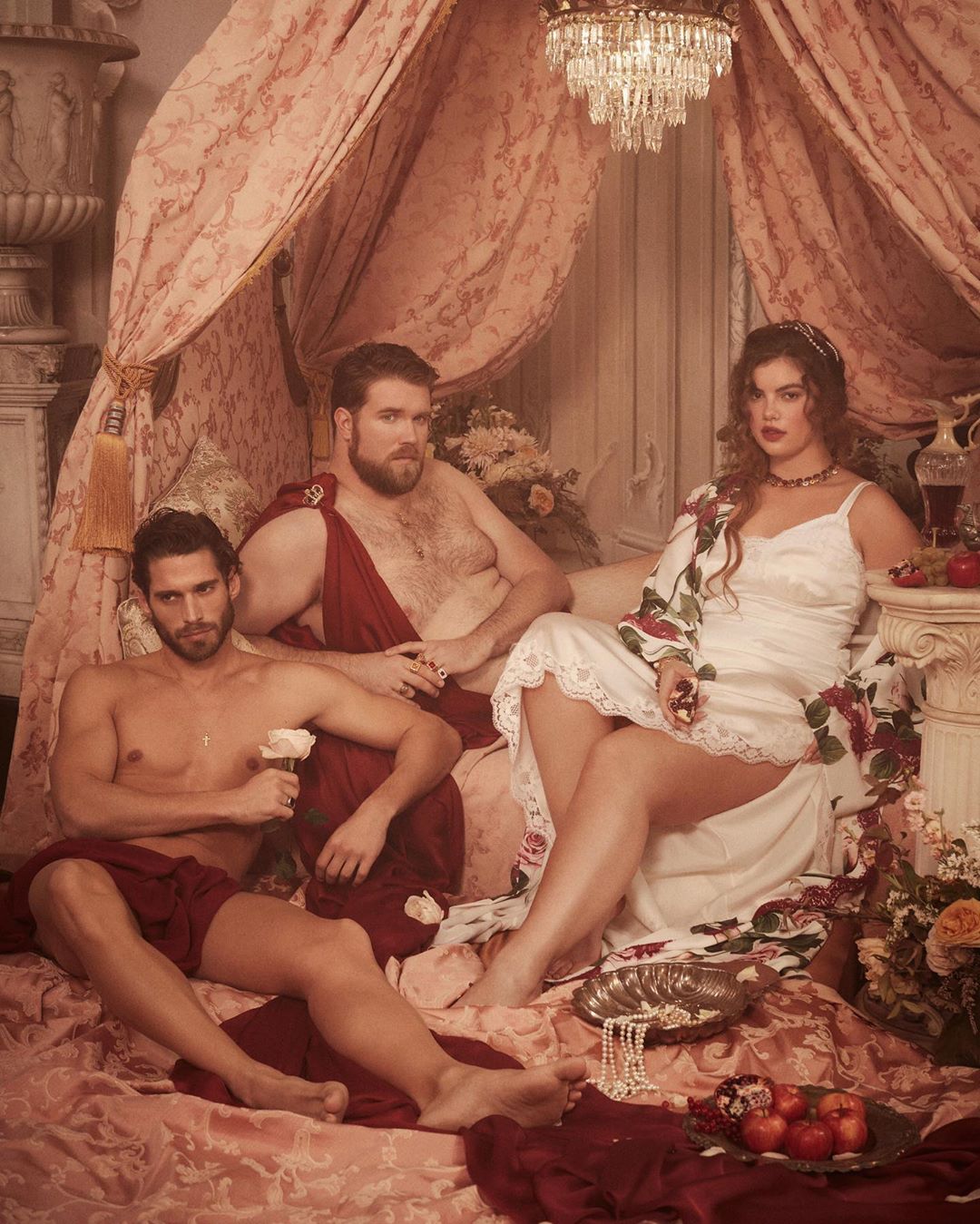 Three people, two men and a woman, in a classical-style photo shoot, draped in fabric, on a bed with classical-era-style decor.