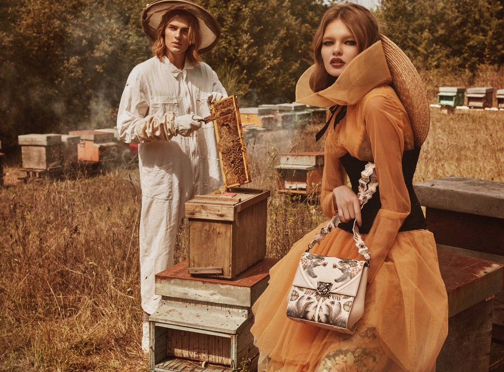 Two models in bee-inspired fashion next to stacked beehives in a field with trees in the background, evoking a vintage editorial style.