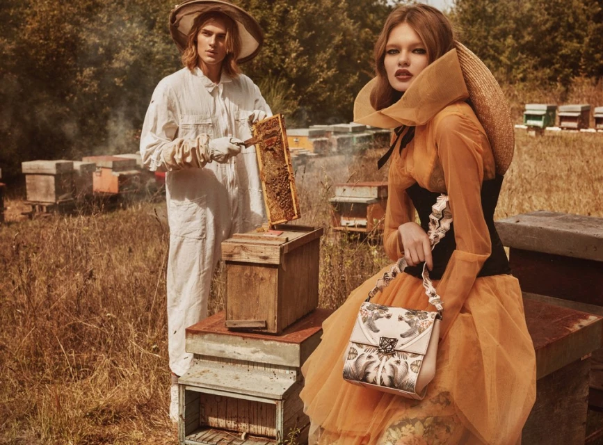 Two models in bee-inspired fashion next to stacked beehives in a field with trees in the background, evoking a vintage editorial style.