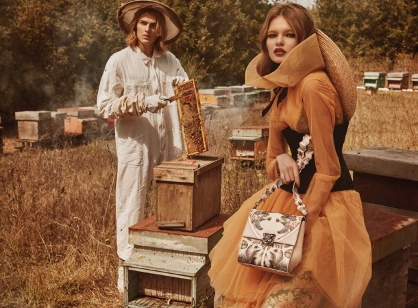 Two models in bee-inspired fashion next to stacked beehives in a field with trees in the background, evoking a vintage editorial style.