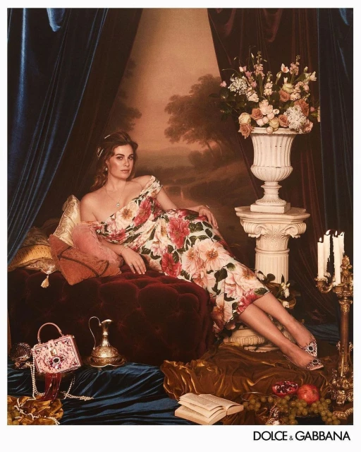 Fashion shot of a woman reclining on a velvet chaise lounge in a floral gown, with a handbag, shoes, and flowers in the foreground.