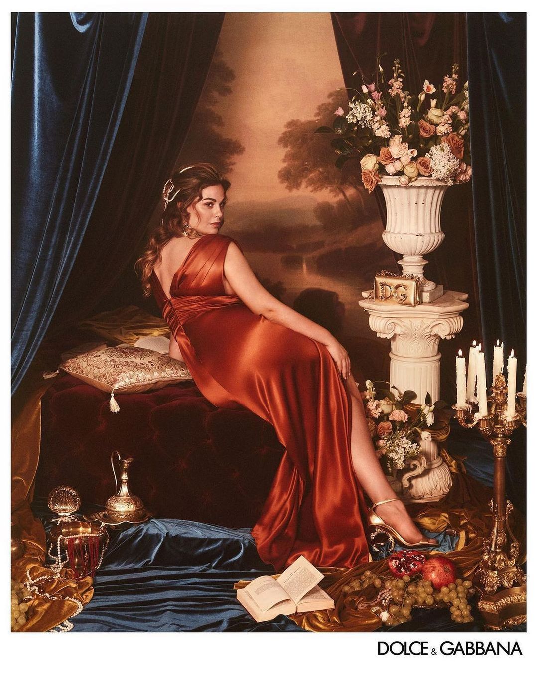Brunette woman in a rust-colored dress poses reclining on a bed with a classical-style still-life background in a studio setting.
