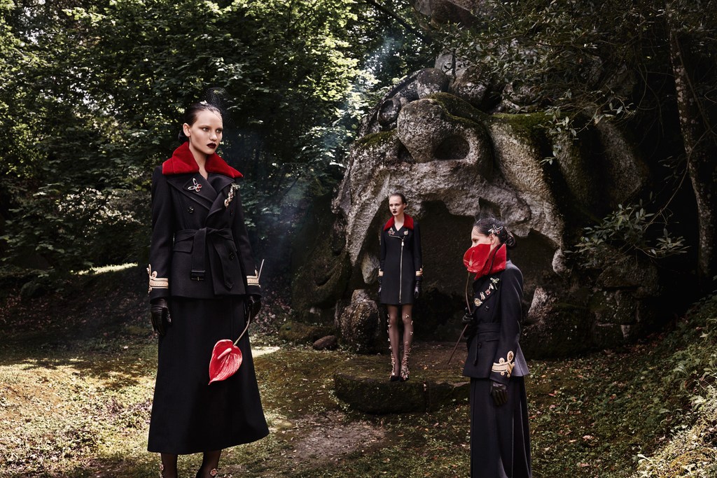 Three women in black coats with red accents stand in a forest clearing near a stone sculpture. Dark, editorial fashion shot.