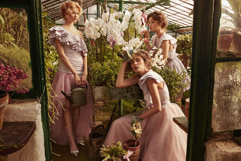 Three women in ethereal dresses pose among orchids and greenery in a sunlit greenhouse. One holds a watering can, exuding a dreamy fashion editorial aesthetic.