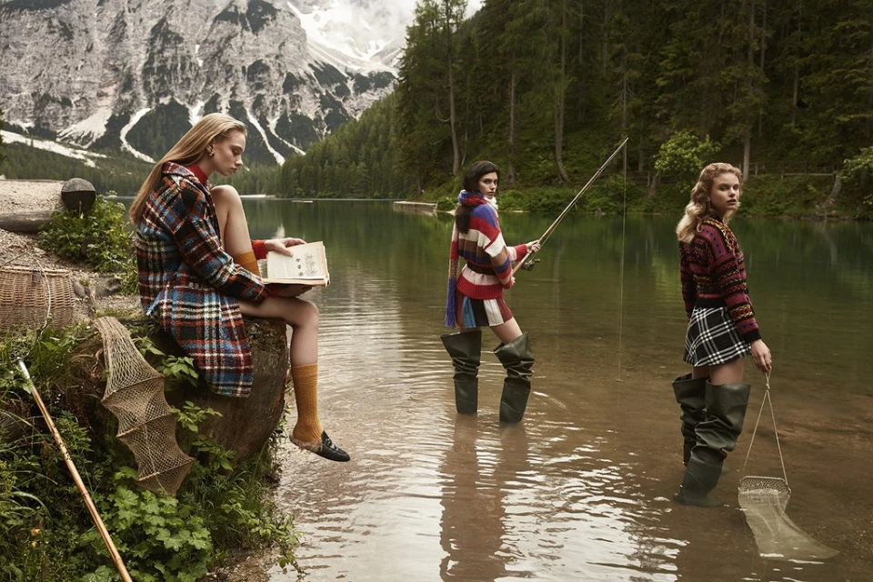Three women stand in a lake with a mountain backdrop. One reads while sitting on a rock, and two are fishing with nets and a fishing pole.