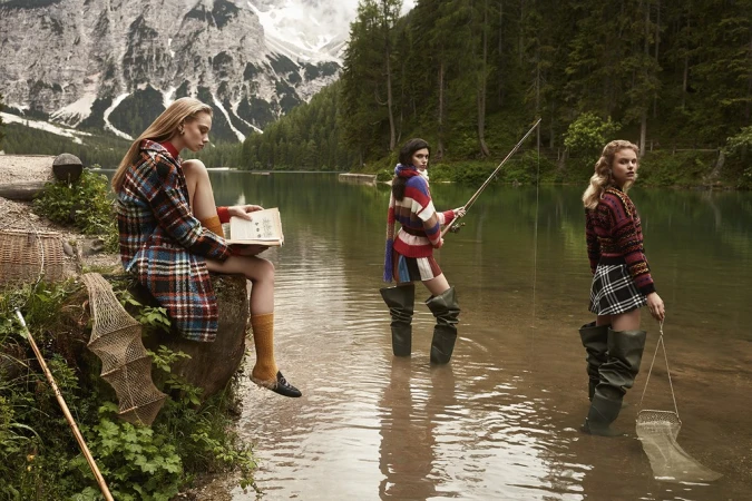 Three women stand in a lake with a mountain backdrop. One reads while sitting on a rock, and two are fishing with nets and a fishing pole.