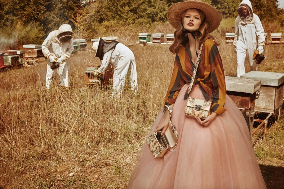 Fashionable woman poses in golden field with beekeepers tending to hives in background, wearing a straw hat and floral purse.