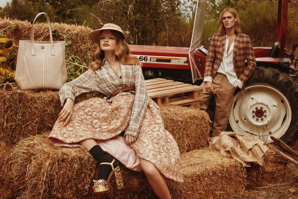 Woman in plaid sitting on hay bales with a handbag and a tractor in the background; man in plaid stands beside a tractor outside.
