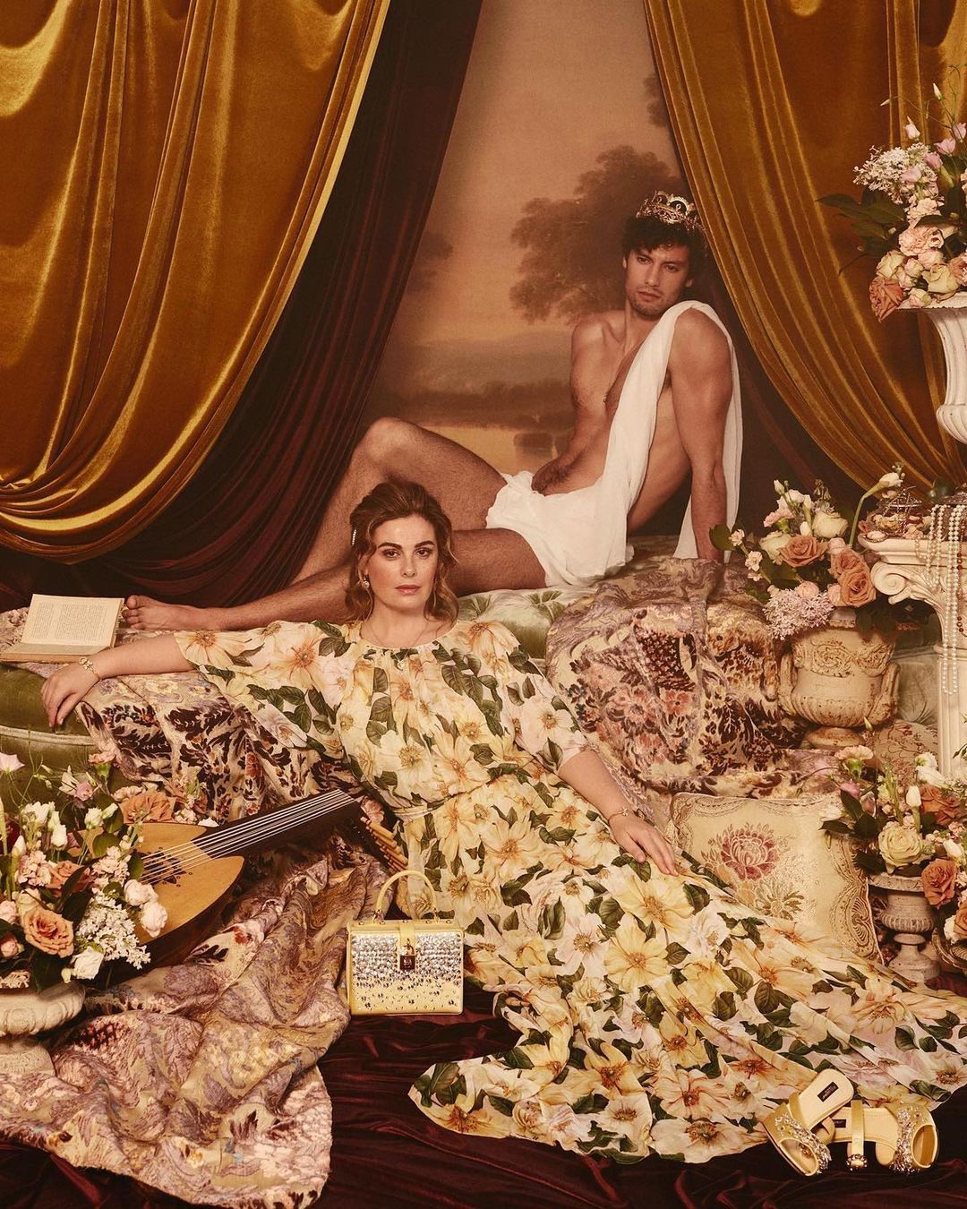 Classical-style image of a woman in a floral dress reclining on a bed with a man wearing a crown and towel draped behind her.