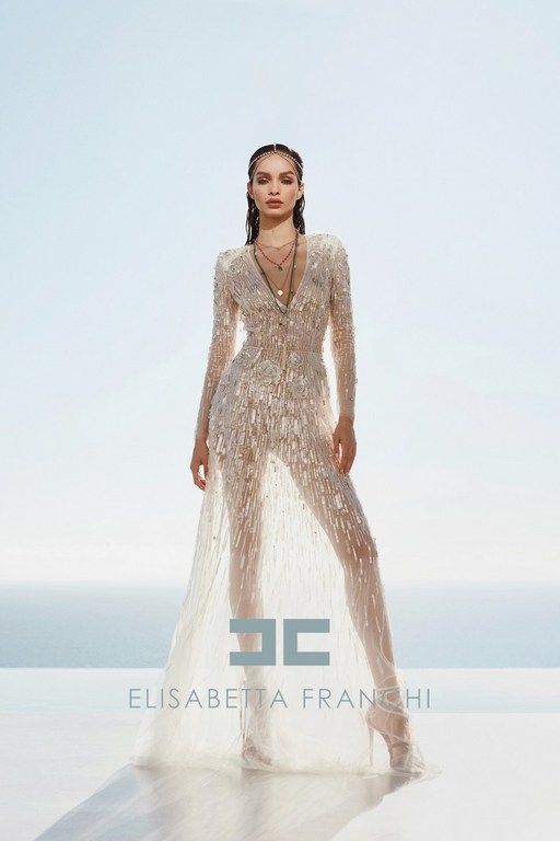 Model in a sheer embellished long-sleeve gown standing against a bright sky backdrop with a brand logo overlay
