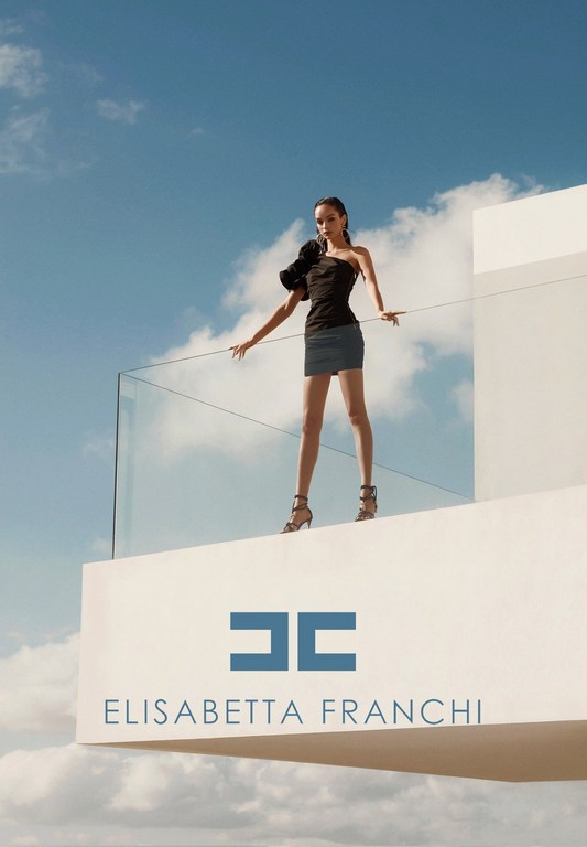 Fashion model in black strapless top and short blue skirt poses on a white rooftop against a blue cloudy sky, promoting Elisabetta Franchi.
