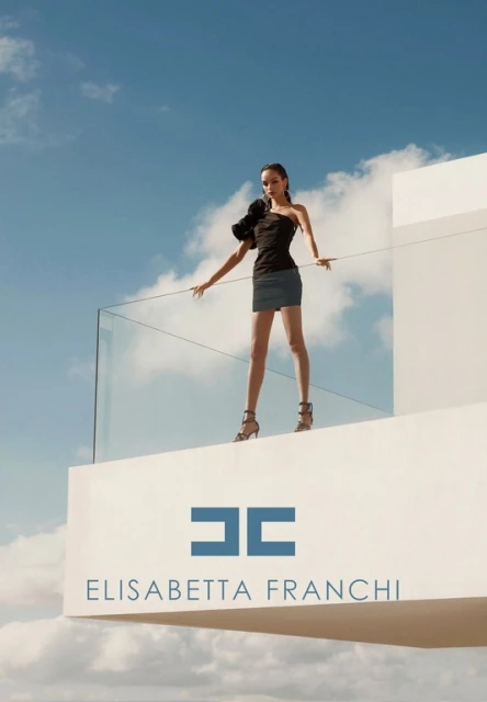 Fashion model in black strapless top and short blue skirt poses on a white rooftop against a blue cloudy sky, promoting Elisabetta Franchi.