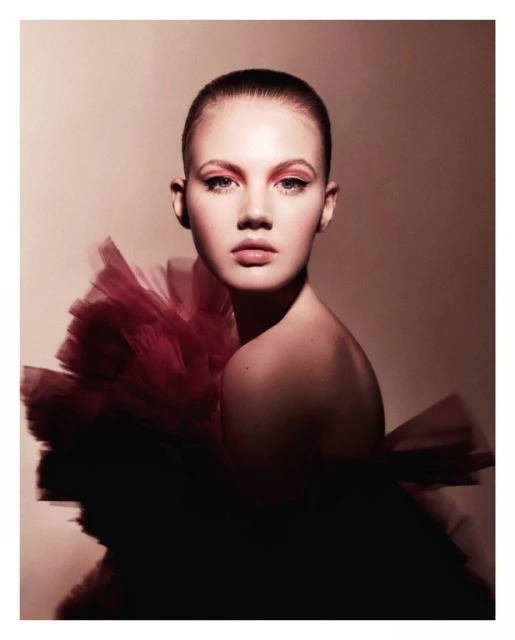 Close-up portrait of a young woman with short hair and red eye makeup wearing a large red tulle dress against a neutral background.