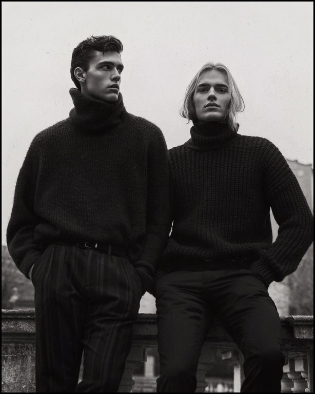 Two male models in dark turtleneck sweaters standing together against an outdoor architectural background in Milan.