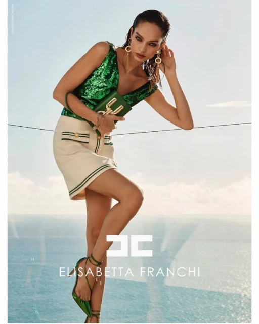 Brunette fashion model poses in a green sequined top and white mini skirt, holding a matching green purse with sky and ocean background.