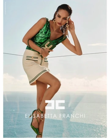Brunette fashion model poses in a green sequined top and white mini skirt, holding a matching green purse with sky and ocean background.