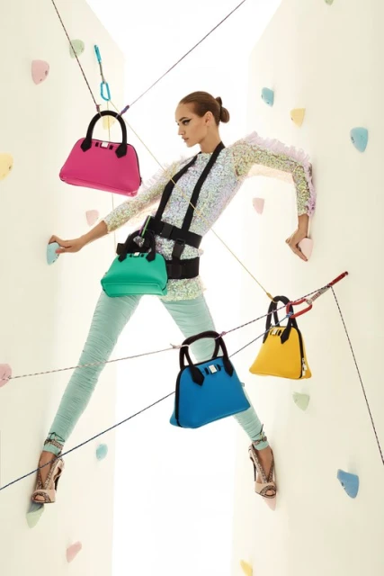 Fashion shot of woman climbing rock wall with colorful handbags attached to climbing ropes, wearing climbing harness and sequin top.