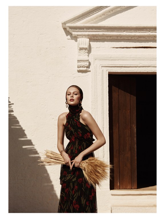 Woman in floral halter dress posing next to a door holding a bundle of wheat in an outdoor setting in natural light.