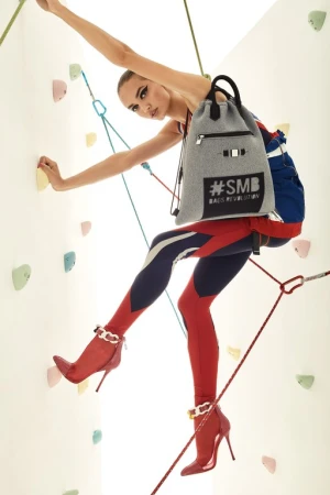 A woman poses on a climbing wall wearing a backpack and red high heels. Colorful climbing holds and ropes are visible in the background.