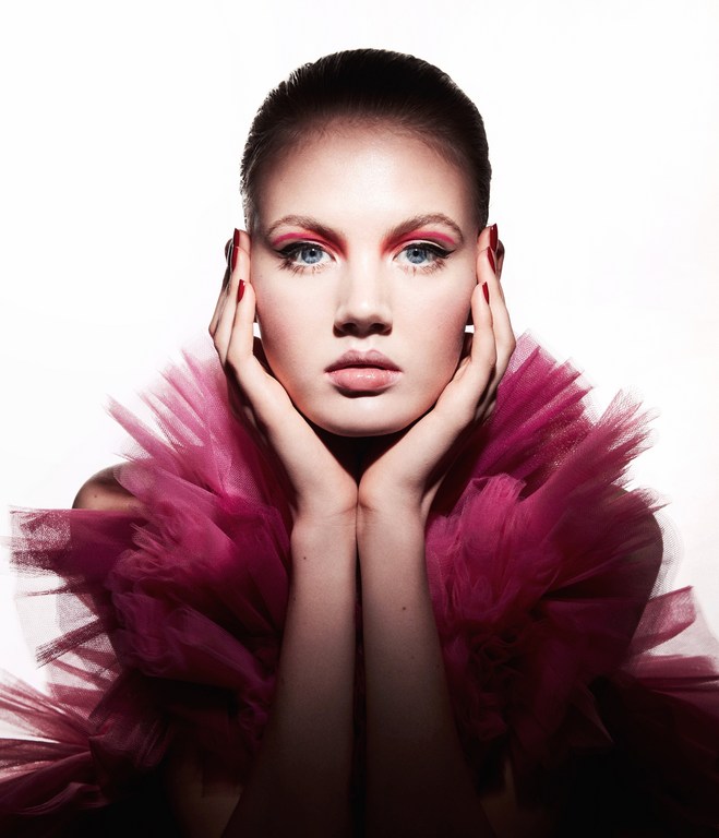 Close-up portrait of a young adult female model with pink eye makeup, full lips, and a pink tulle ruffled collar on a white background.